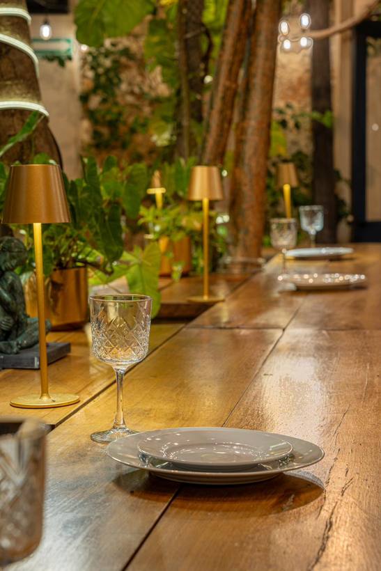 Mesa larga de madera con lámparas doradas, copas y plantas al fondo